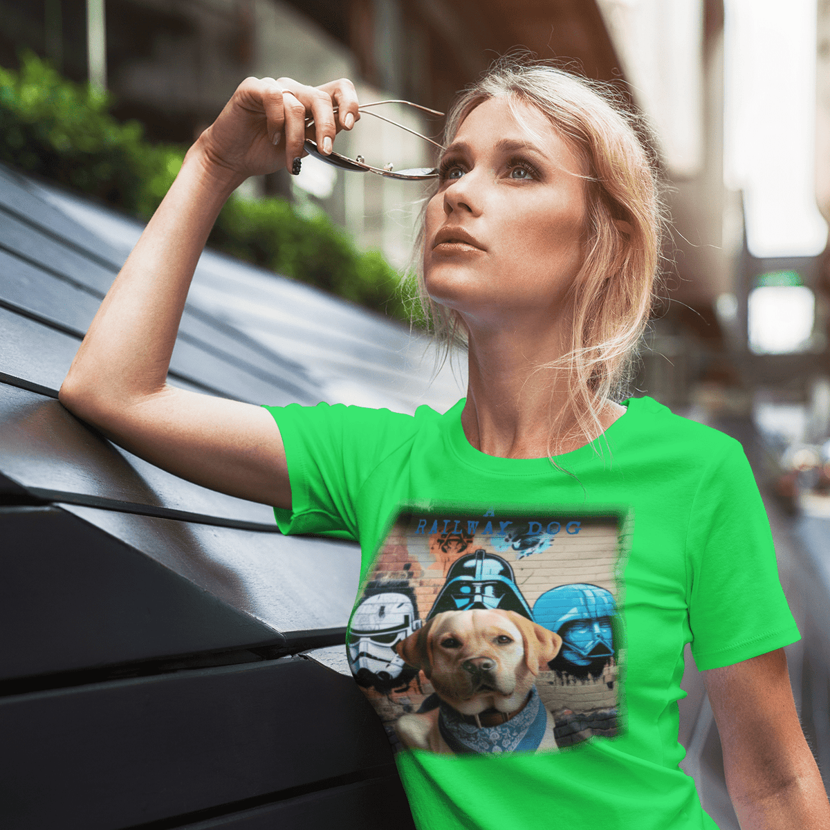 Blonde woman looking upward while wearing a bright neon green t-shirt featuring a Yellow Labrador wearing a bandana in front of a brick wall with sci-fi helmet street art and "A Railway Dog" text.