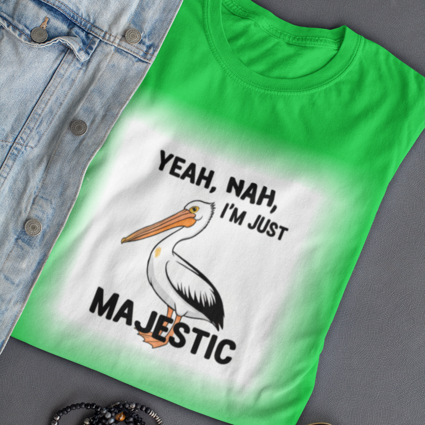 Green t-shirt with a pelican graphic and text, placed on a gray surface with sunglasses and bracelets.