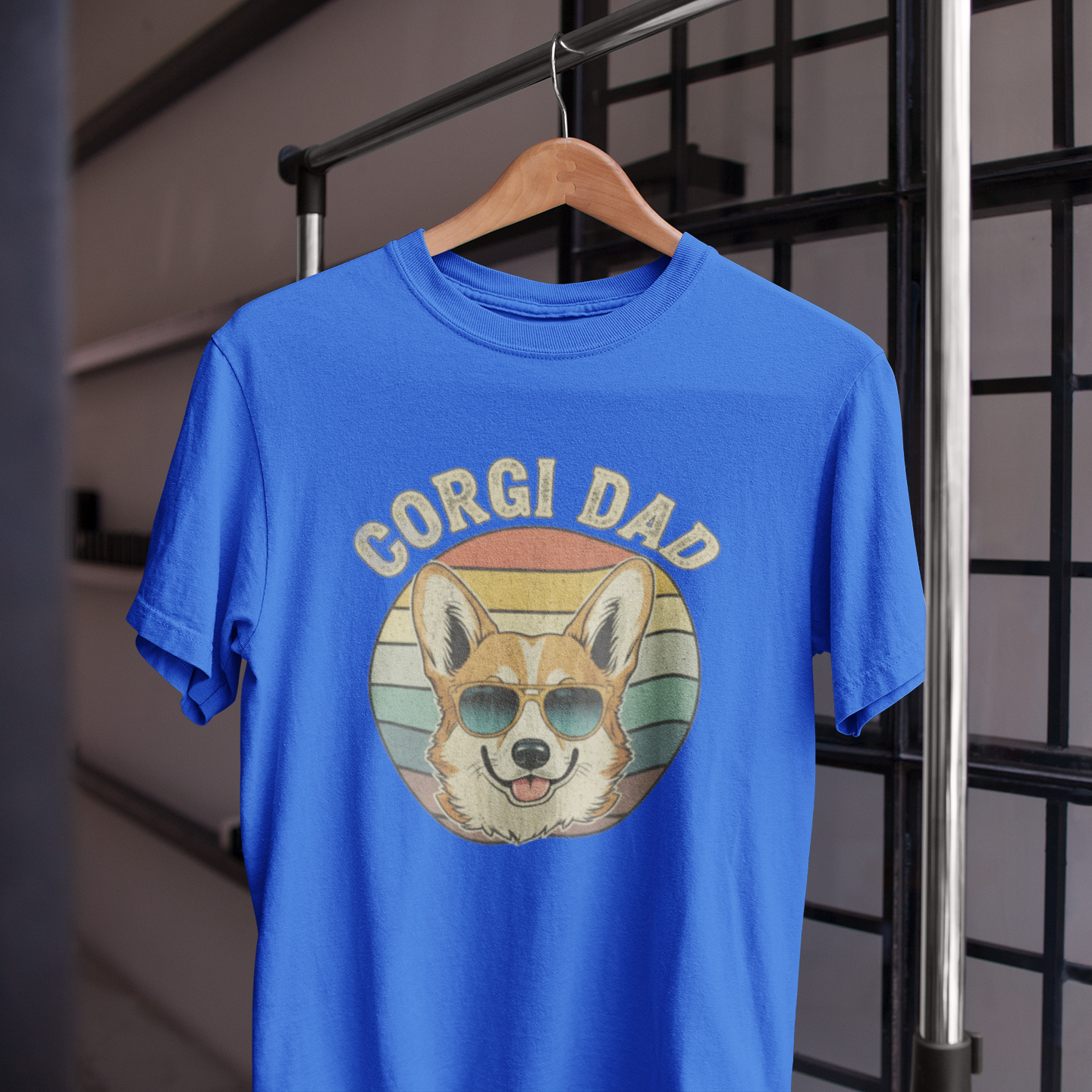 Blue t-shirt with 'Corgi Dad' graphic on a hanger against a metal grid background